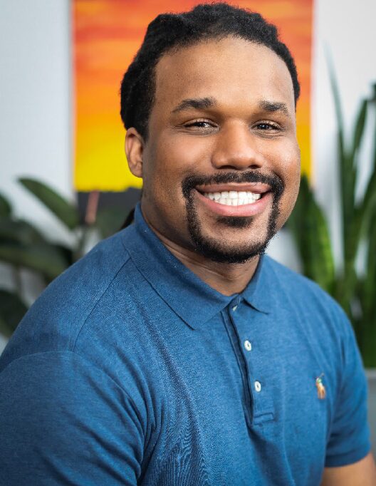 Smiling man with short curly hair and a goatee, wearing a blue polo shirt, in an indoor setting with plants and colorful background.