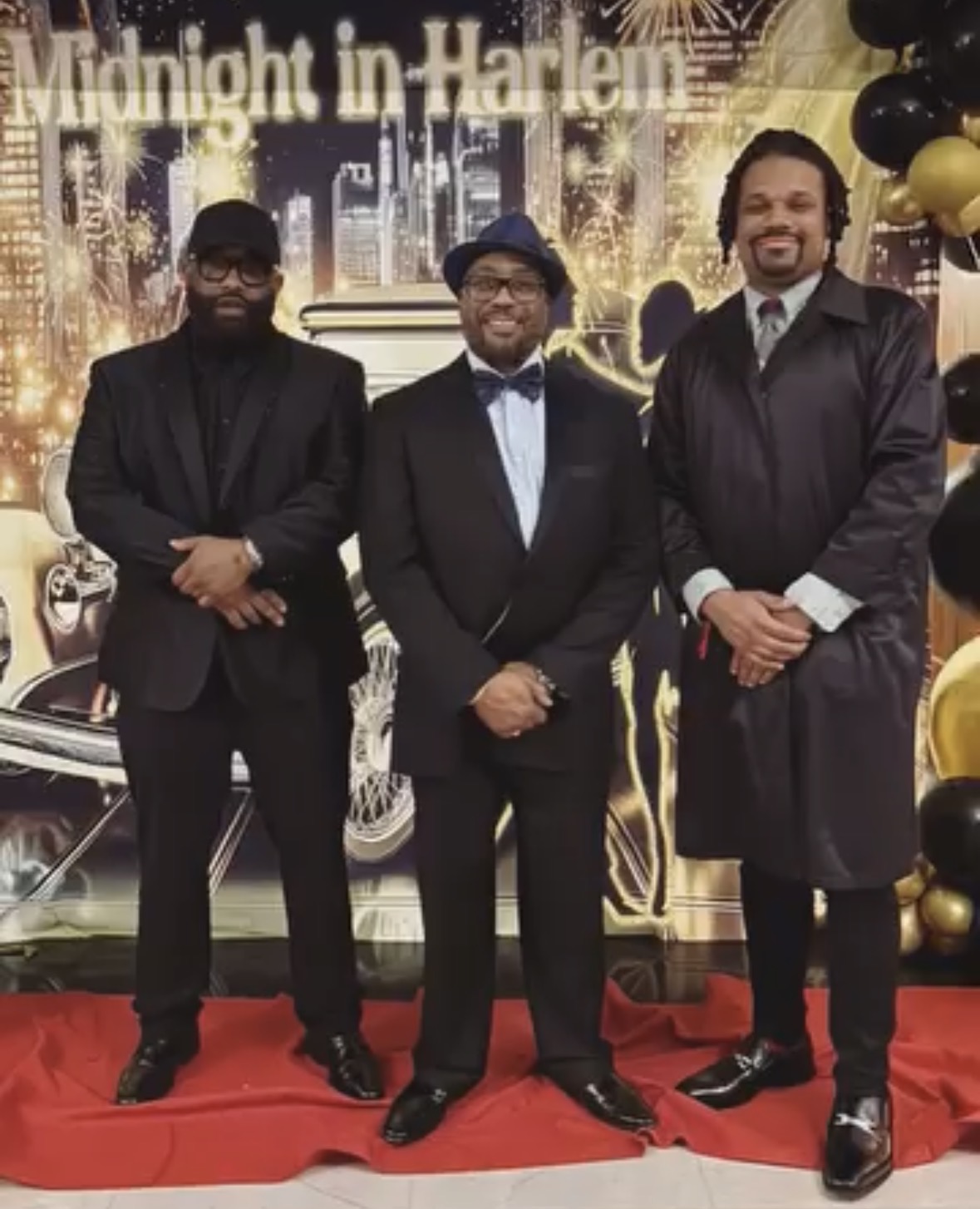 Three men in formal attire standing on a red carpet with a festive backdrop and balloons, smiling at the camera.