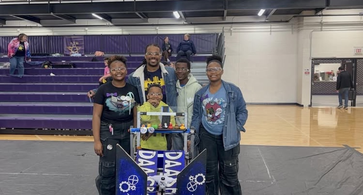 Group of five people standing behind a table with a blue banner, in an indoor gymnasium with purple bleachers.