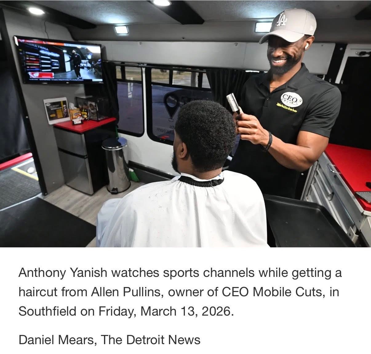 Man getting a haircut while watching sports channels on a screen inside a mobile barbershop, with a customer seated in front.