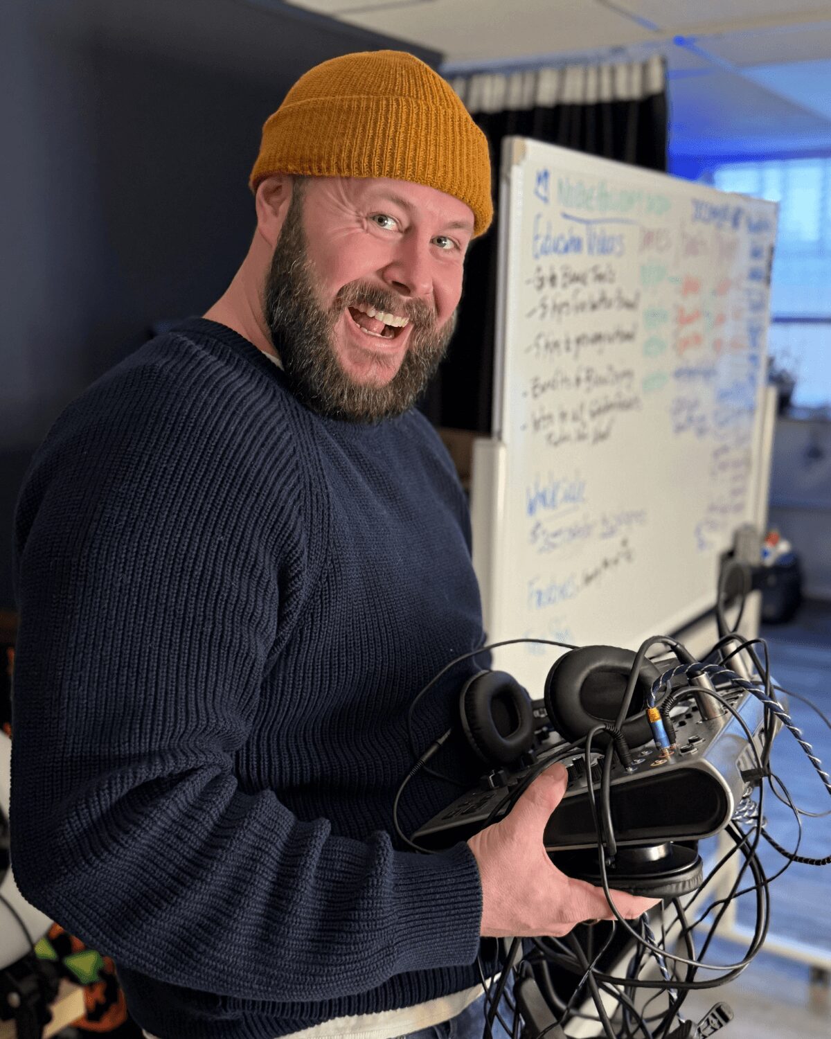 Man with a beard wearing a yellow beanie and dark sweater, smiling, holding headphones and electronic equipment, in a room with a whiteboard.