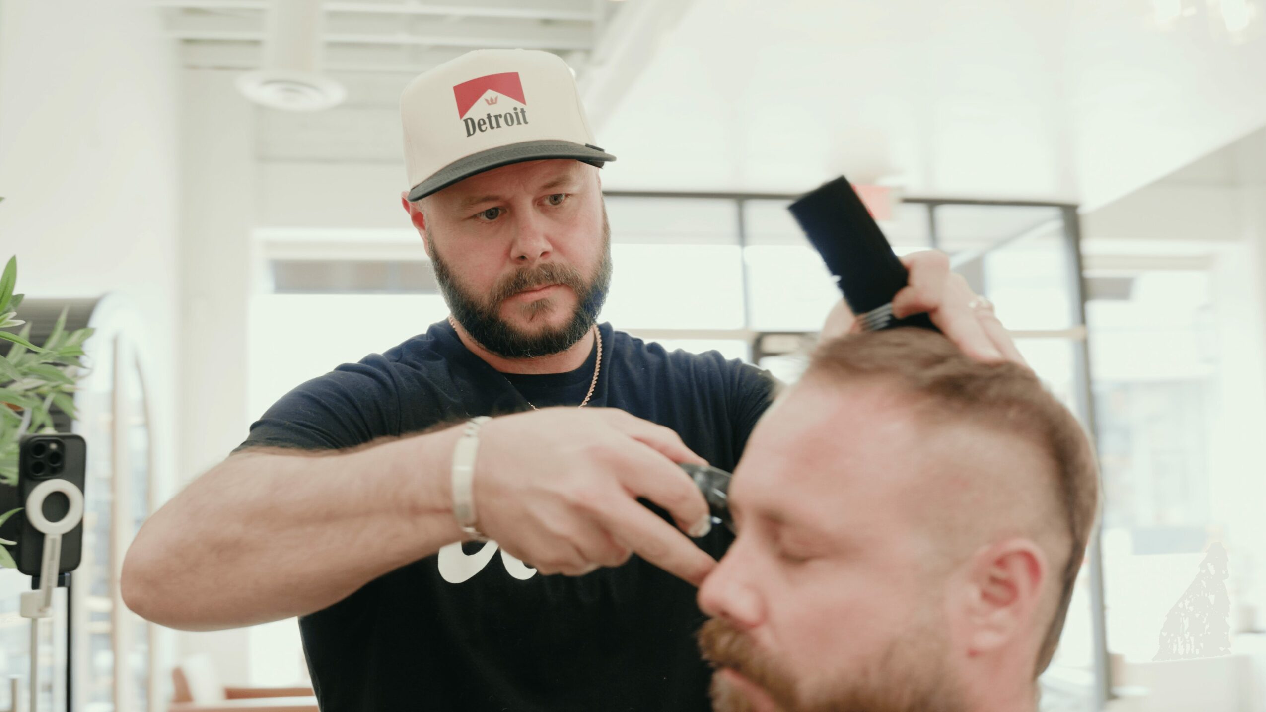 Barber trims a man's hair in a bright, modern salon with large windows and plants.