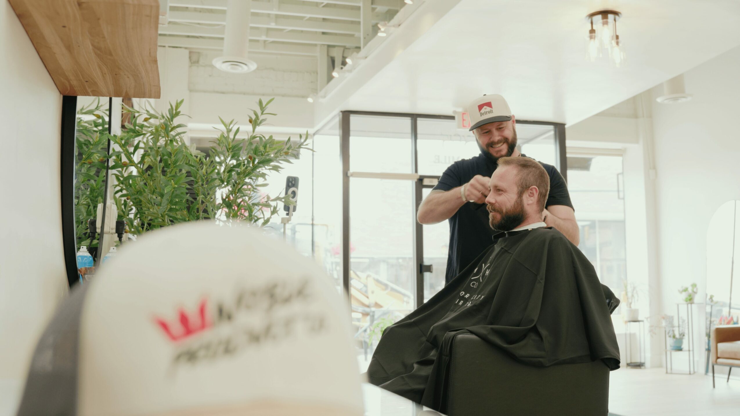 Barber cutting a customer's hair in a bright, modern salon with large windows and greenery.
