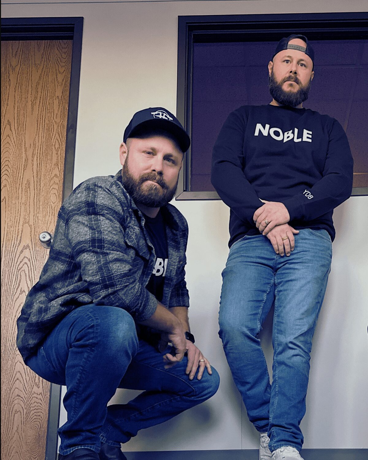 Two men with beards and casual clothing pose indoors, one crouching and one standing, in front of a window and door.