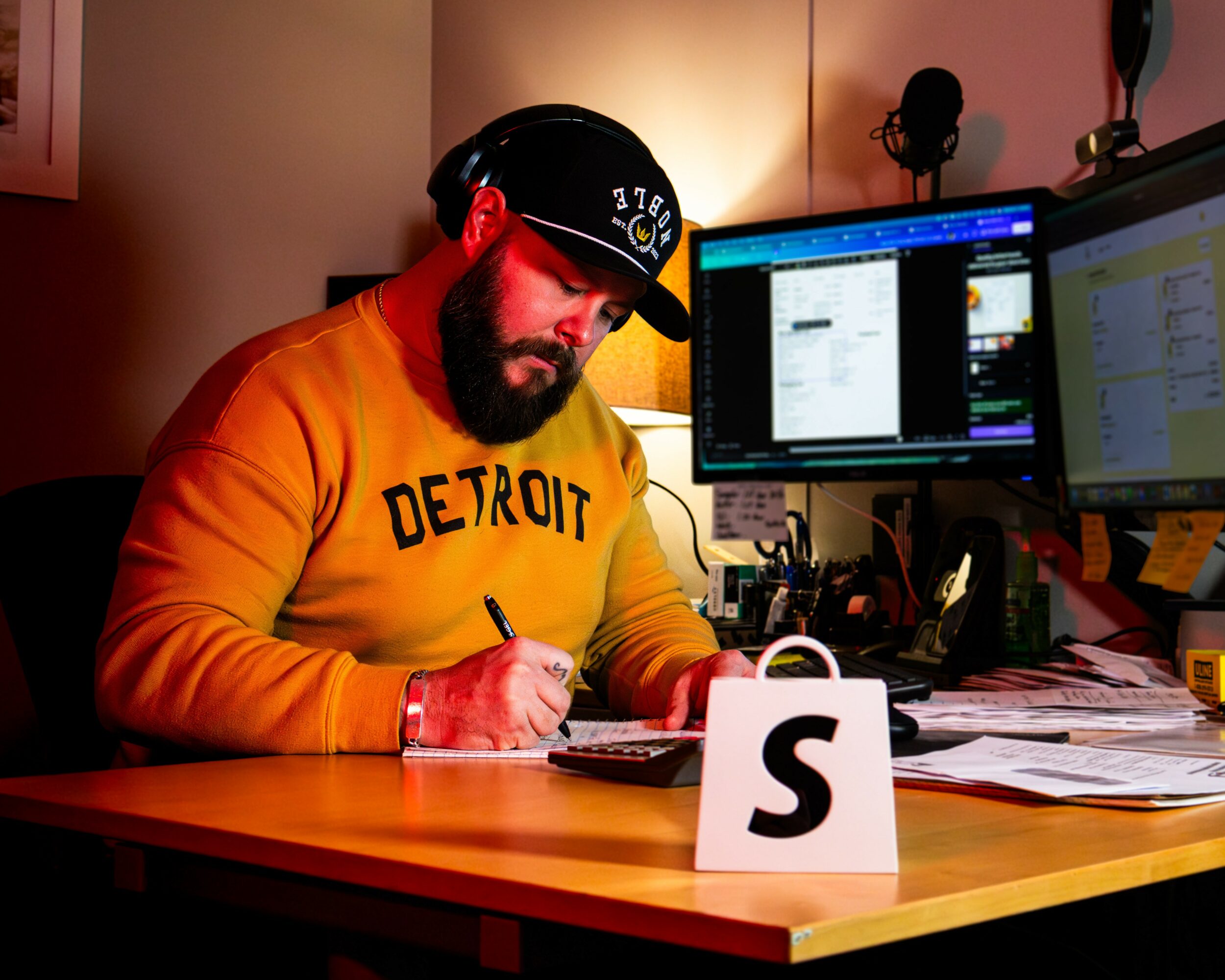 Man with a beard wearing a black cap and orange sweatshirt working at a desk with multiple monitors and papers.