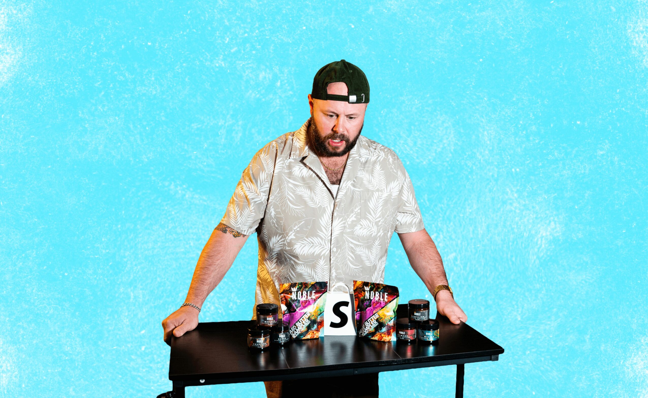 Man with a beard wearing a cap backwards stands behind a table with colorful objects, against a blue background.