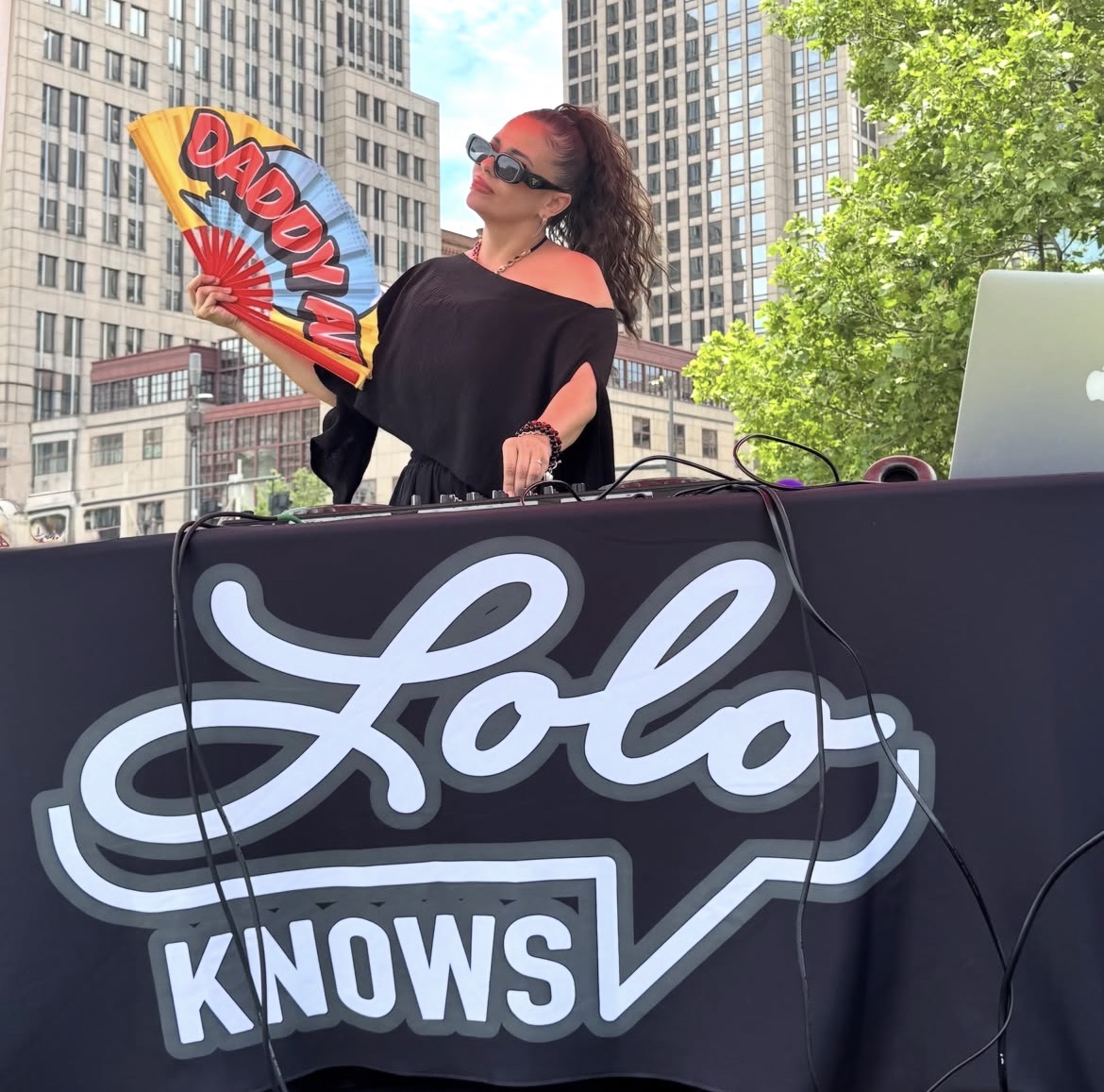 Woman with sunglasses and dark clothing standing behind a DJ booth outdoors, city buildings and trees in background.