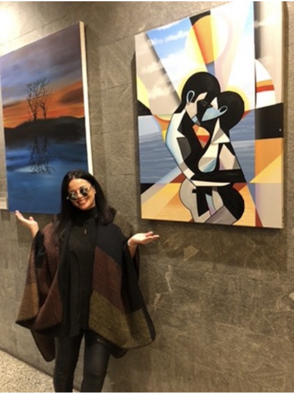 Person with glasses standing in front of colorful abstract paintings on a wall, smiling with arms outstretched.