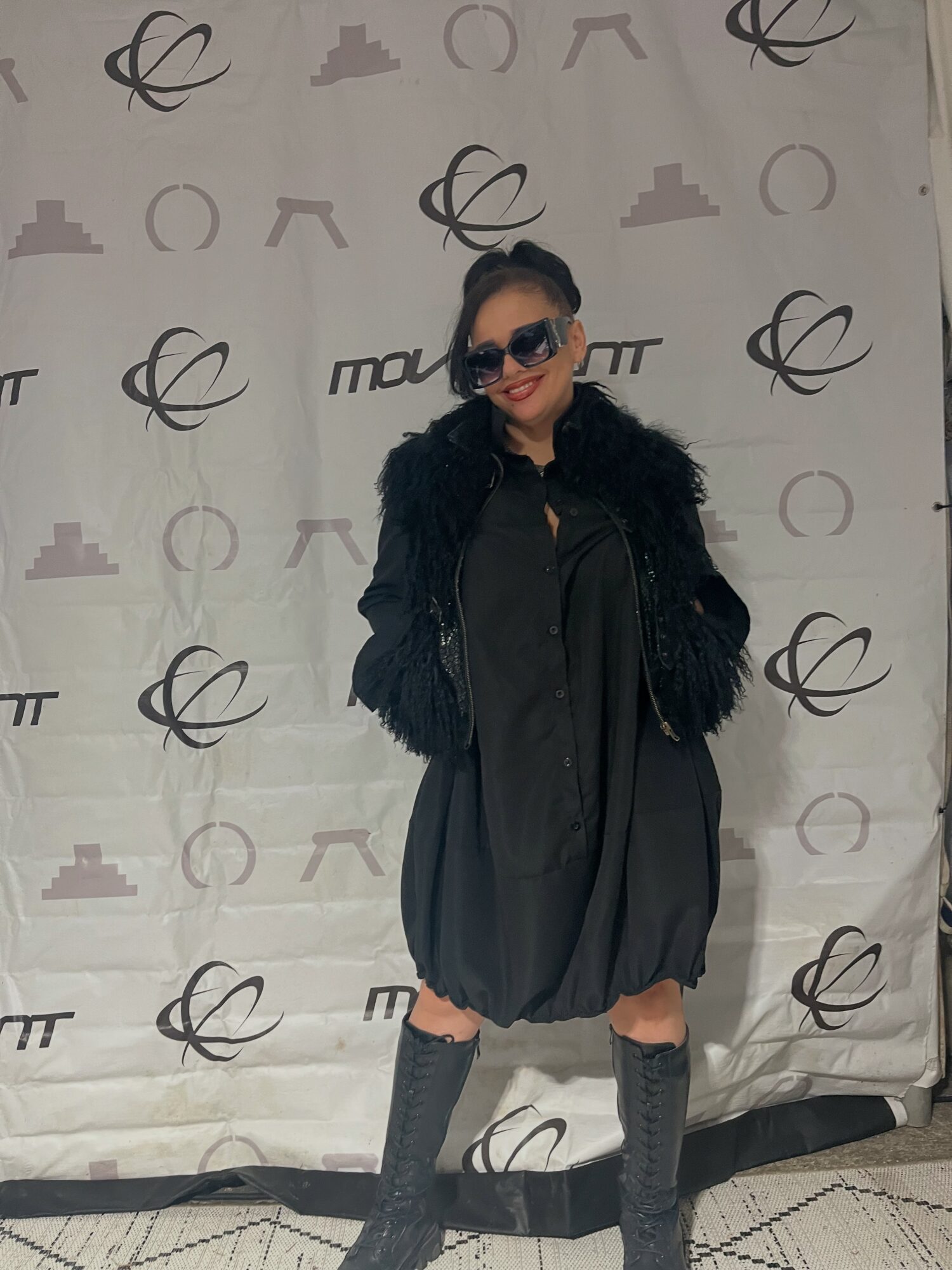 Woman wearing sunglasses, black jacket, and knee-high socks, standing in front of a patterned backdrop.