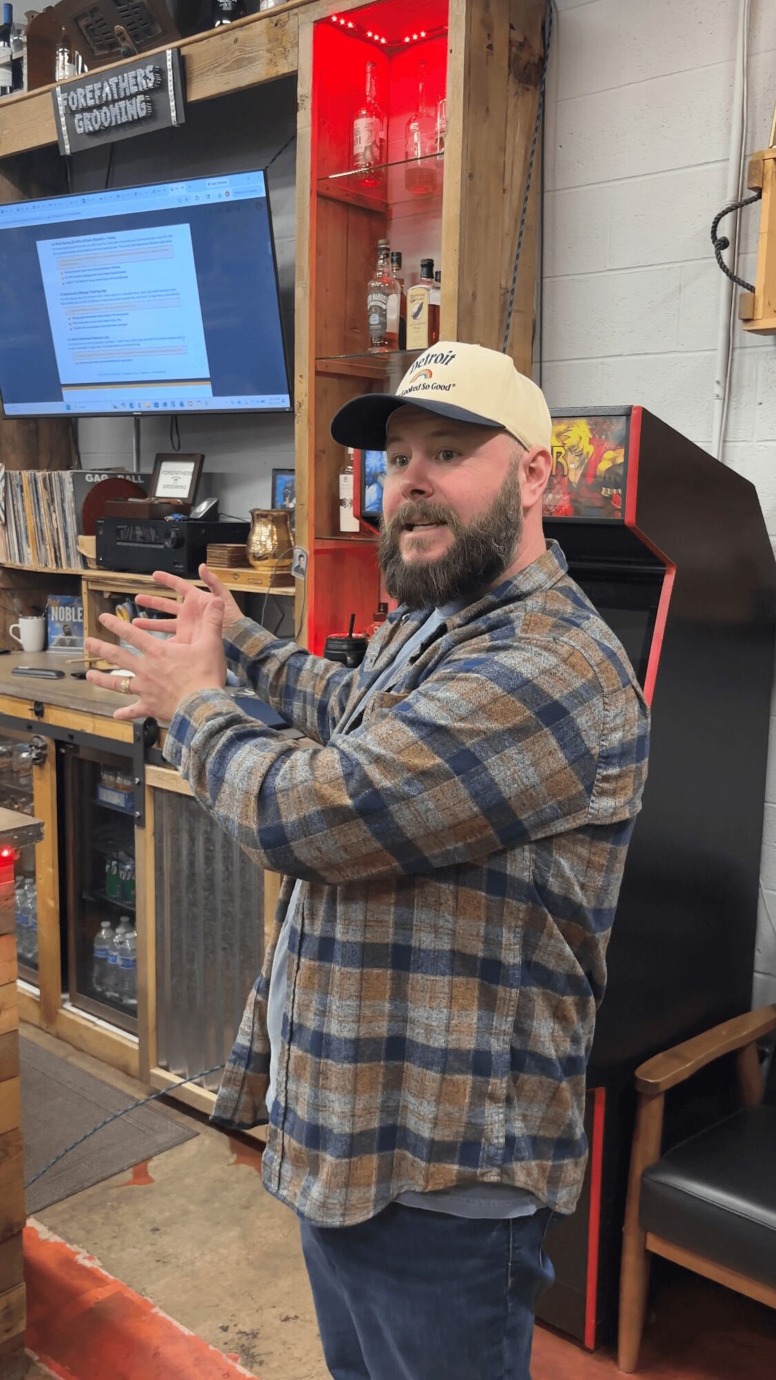 Man with a beard wearing a beige cap and plaid shirt gesturing with hands indoors, shelves and a screen behind him.