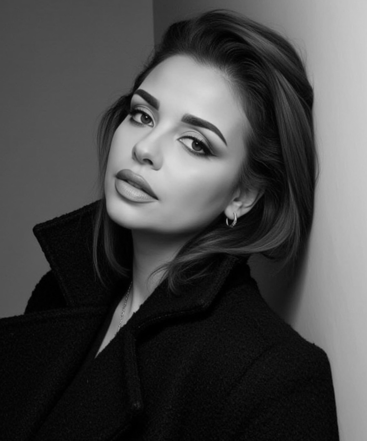 Black and white portrait of a woman with styled hair, makeup, and a serious expression, leaning against a wall.