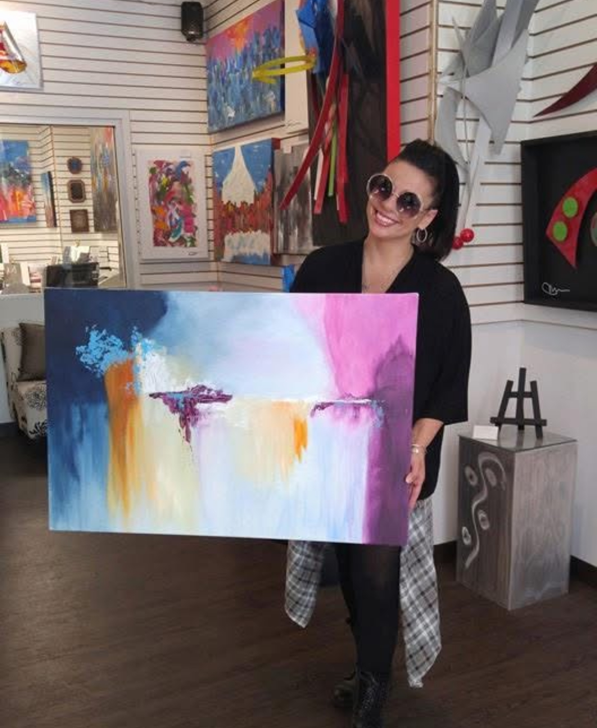 Woman holding colorful abstract painting in art gallery, smiling, wearing sunglasses, black top, plaid shirt tied around waist.