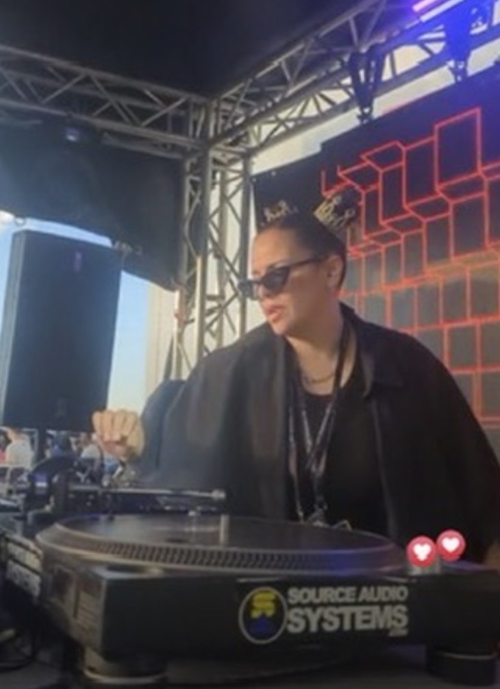 Person wearing sunglasses and a black jacket operating DJ equipment on stage with a geometric background.