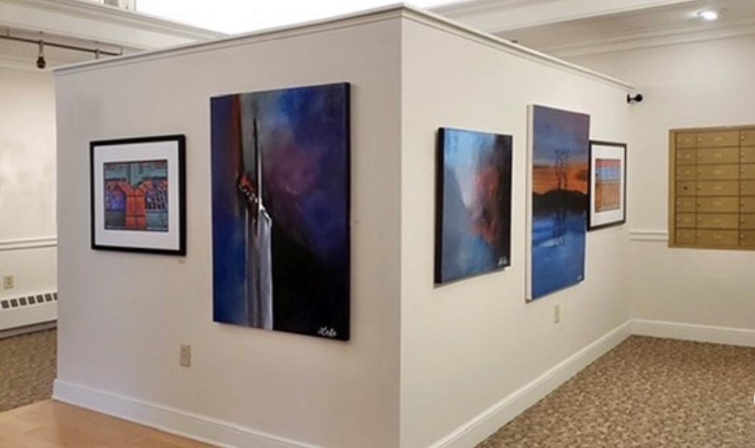 Gallery wall with five abstract paintings on white walls, carpeted floor, and a wooden cabinet on the right.