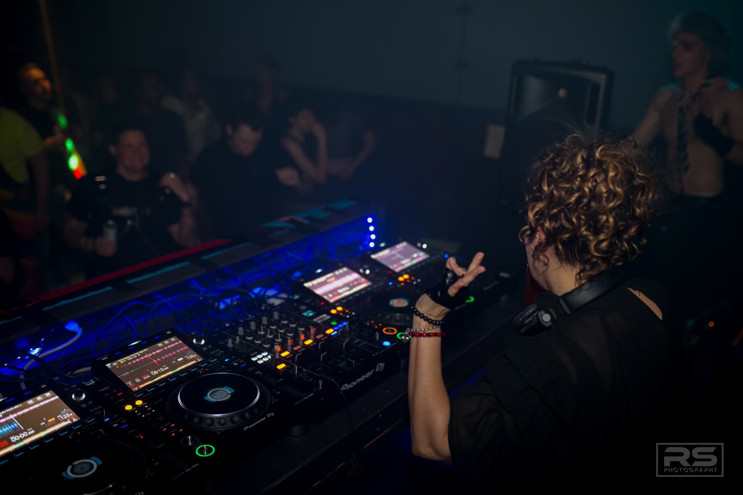 DJ with curly hair performing at a club with a crowd in the background, illuminated by colorful lights.