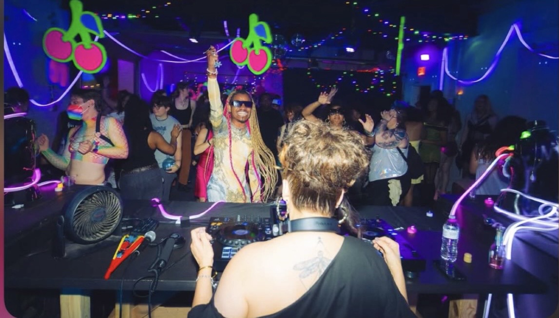 People dancing in a nightclub with colorful lights and decorations, DJ in foreground, lively atmosphere.