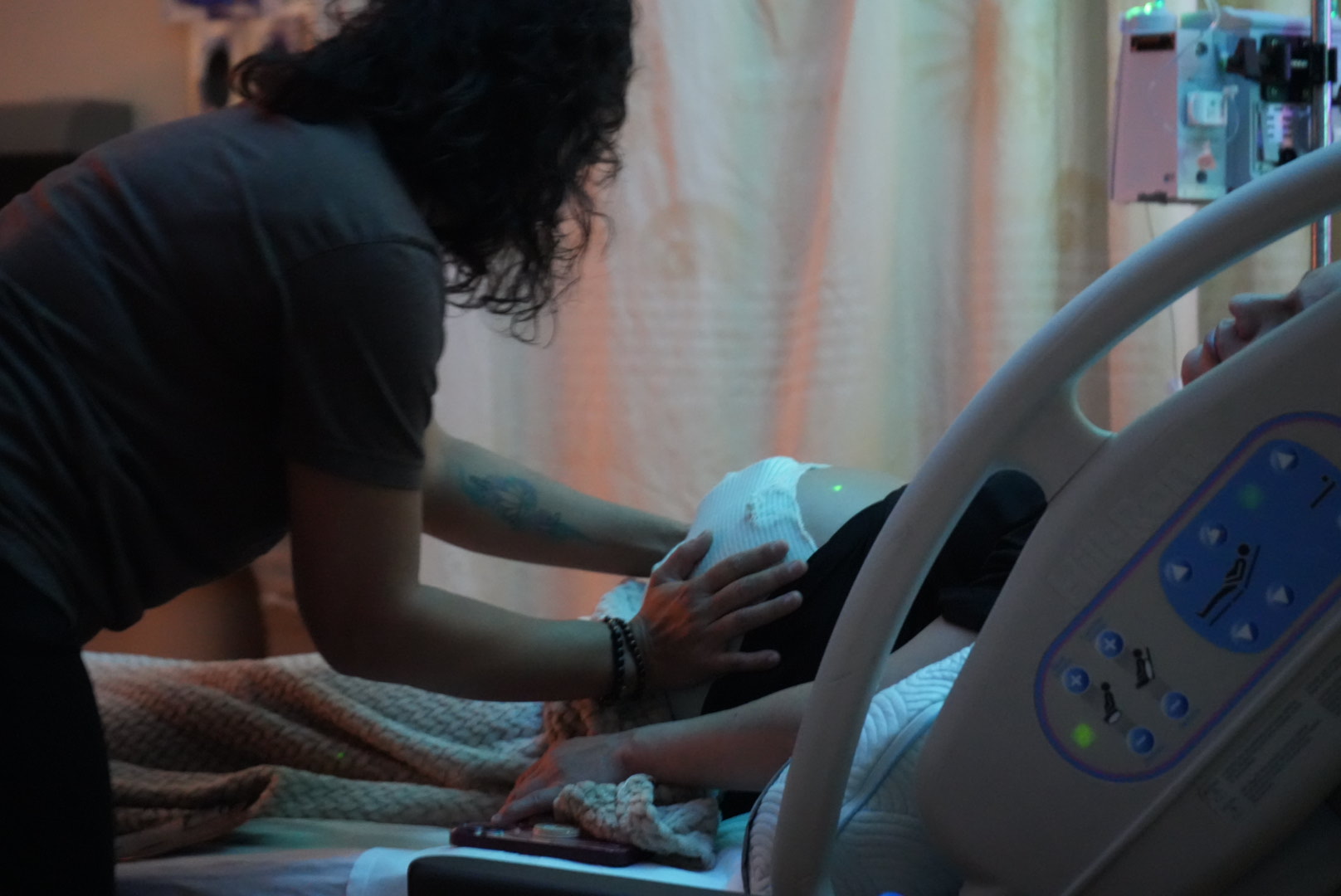 Person leaning over a hospital bed, holding a medical device, with medical equipment in the background.