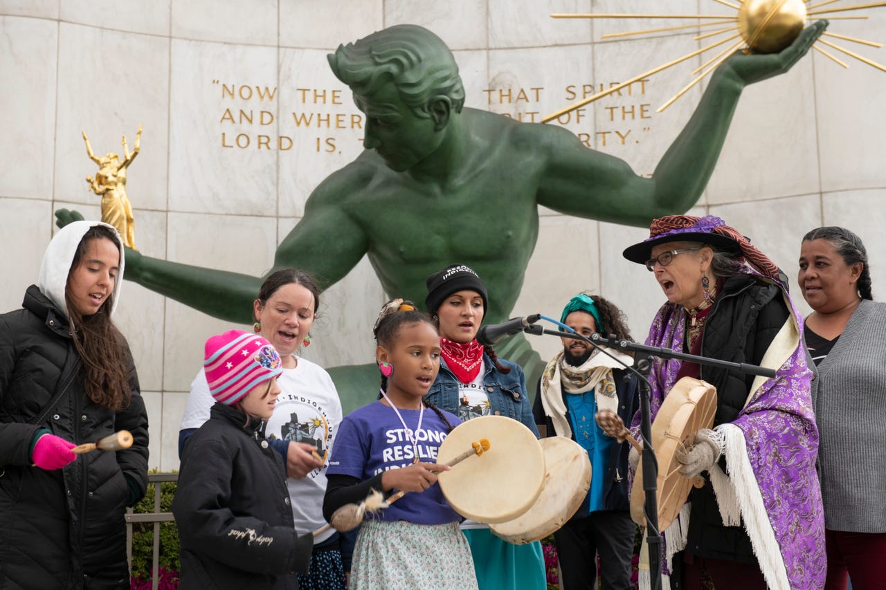 Group of children and adults gathered around a large green statue of a man with outstretched arms, holding a globe, in front of a wall with text.
