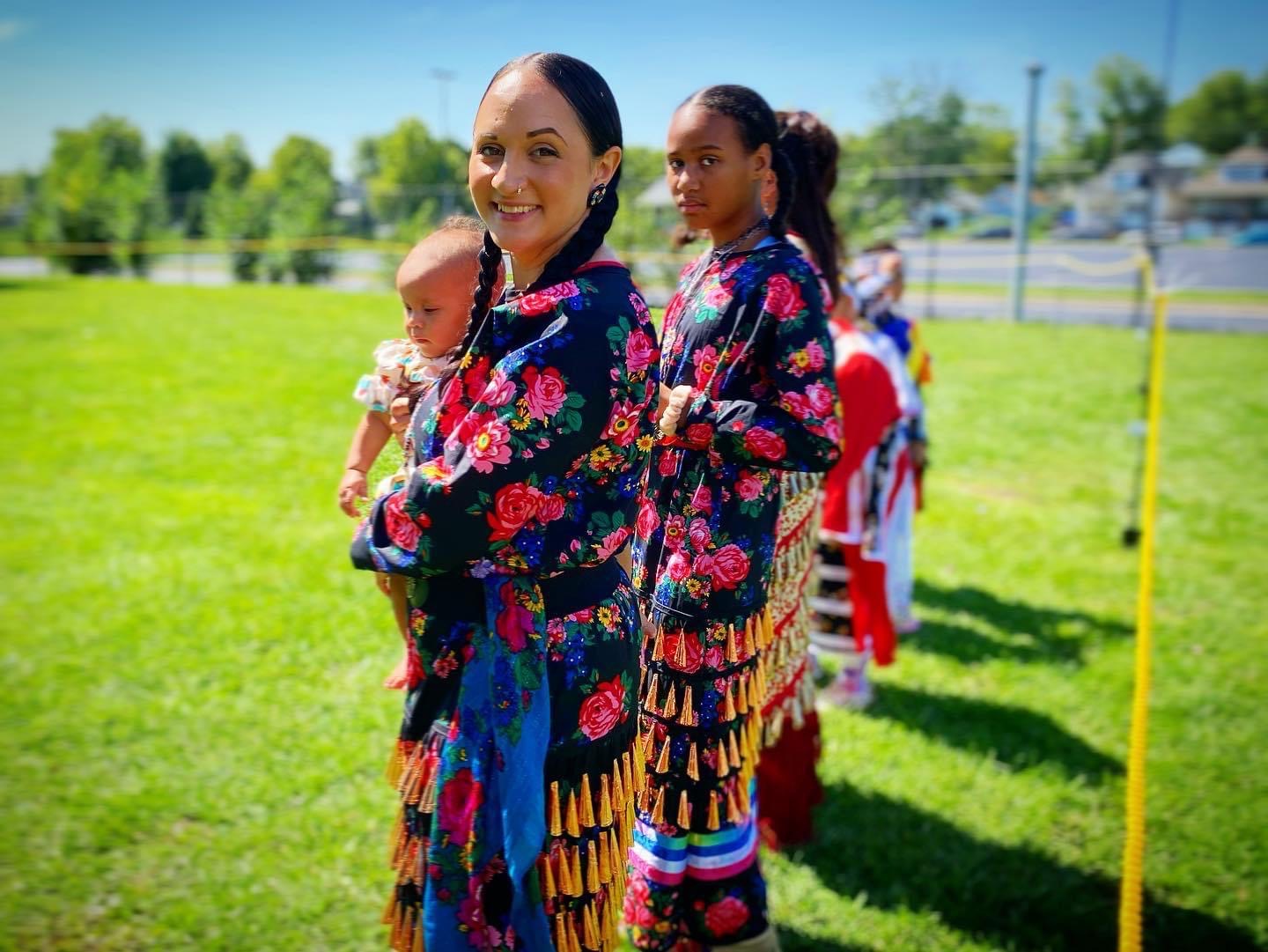 Group of women and children outdoors on grass, wearing colorful traditional clothing, smiling, with trees and a fence in background.