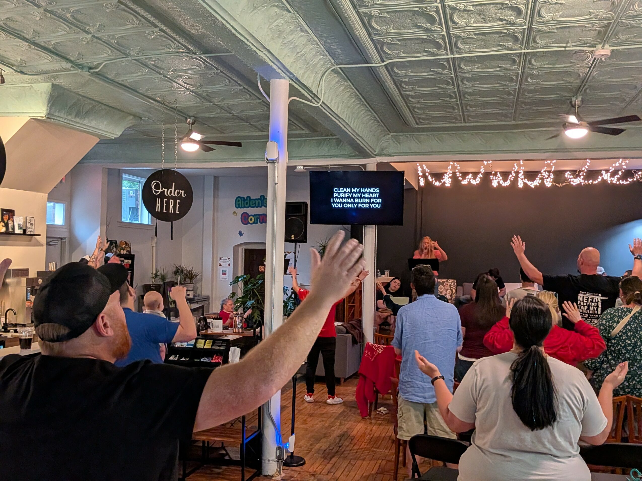 People gathered in a room watching a stage with a speaker, some raising hands, decorated with flowers and string lights.