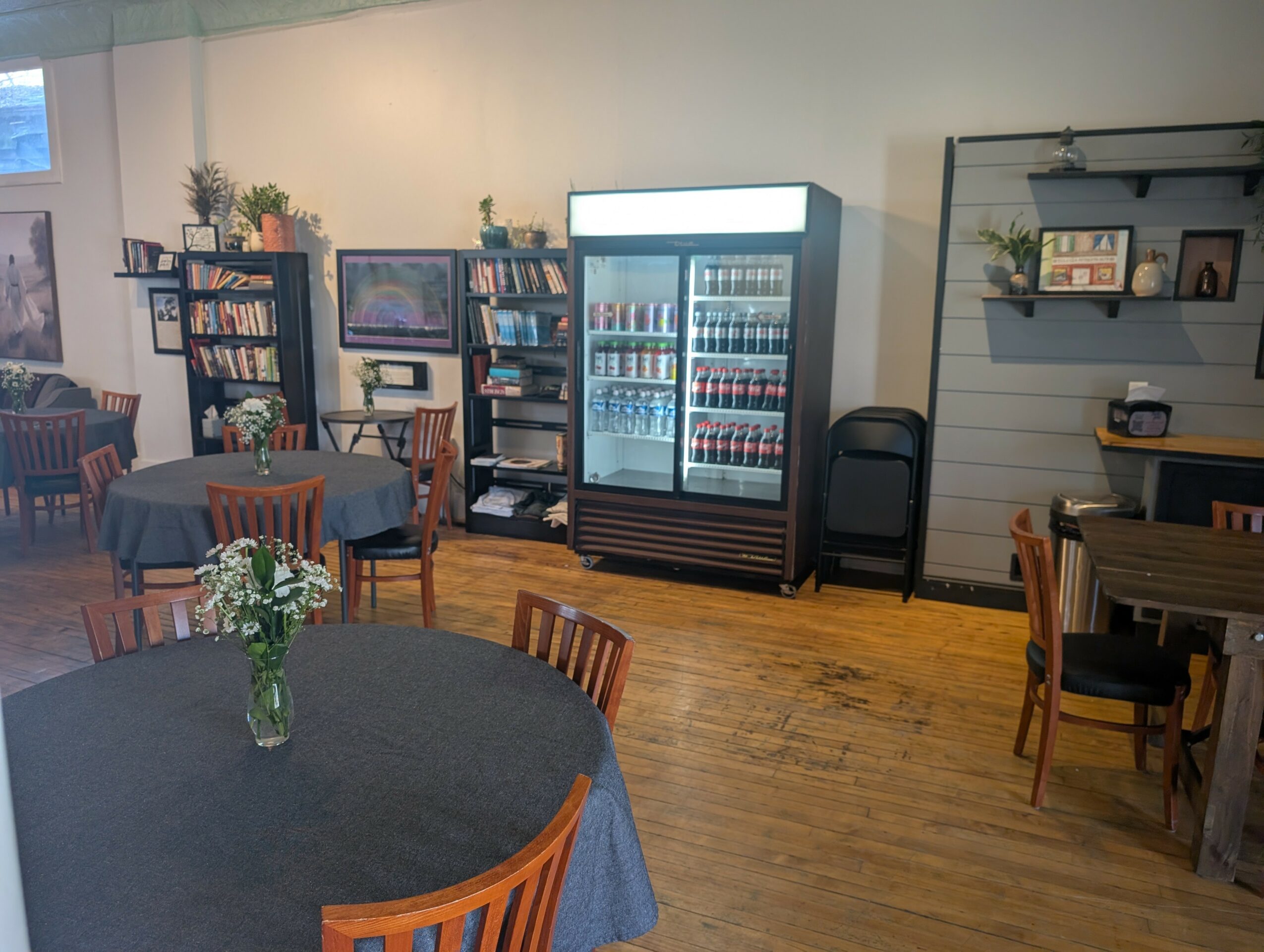 Room with round table, chairs, bookshelf, and a beverage fridge, decorated with plants and framed pictures, wooden floor.