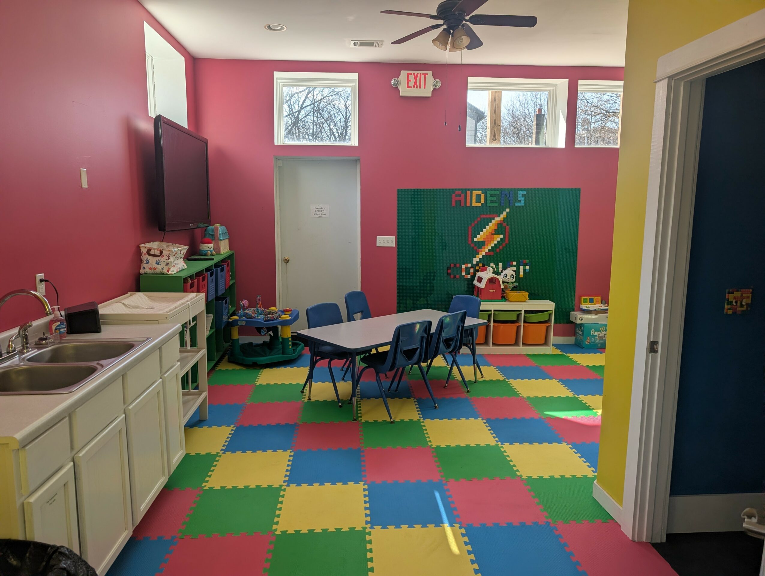 Colorful classroom with a table, chairs, a chalkboard, and a play area with foam tiles, bright walls, and windows.