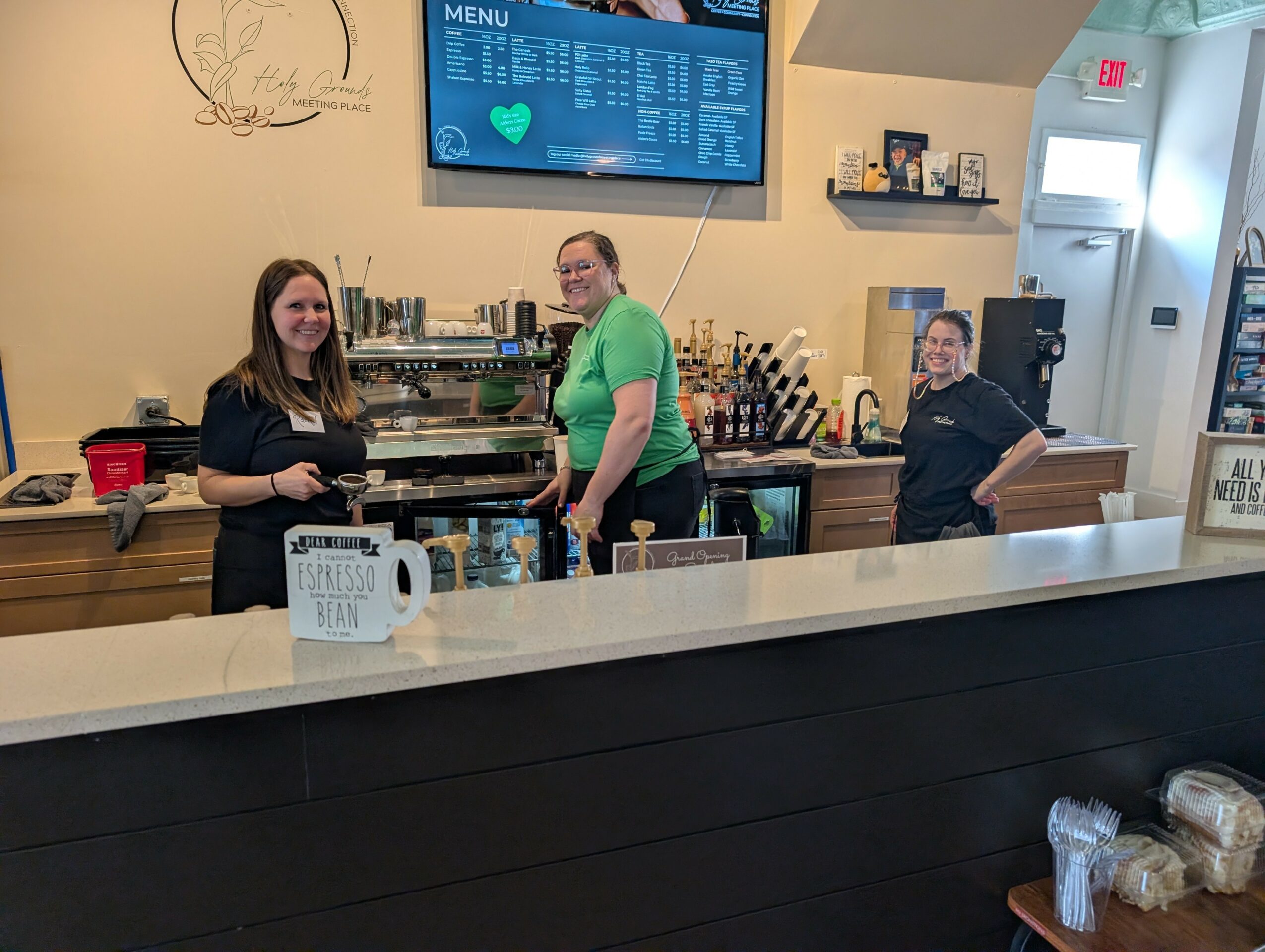 Three women behind a bar counter in a cafe, smiling, with a menu screen above them, and various bottles and items on the counter.