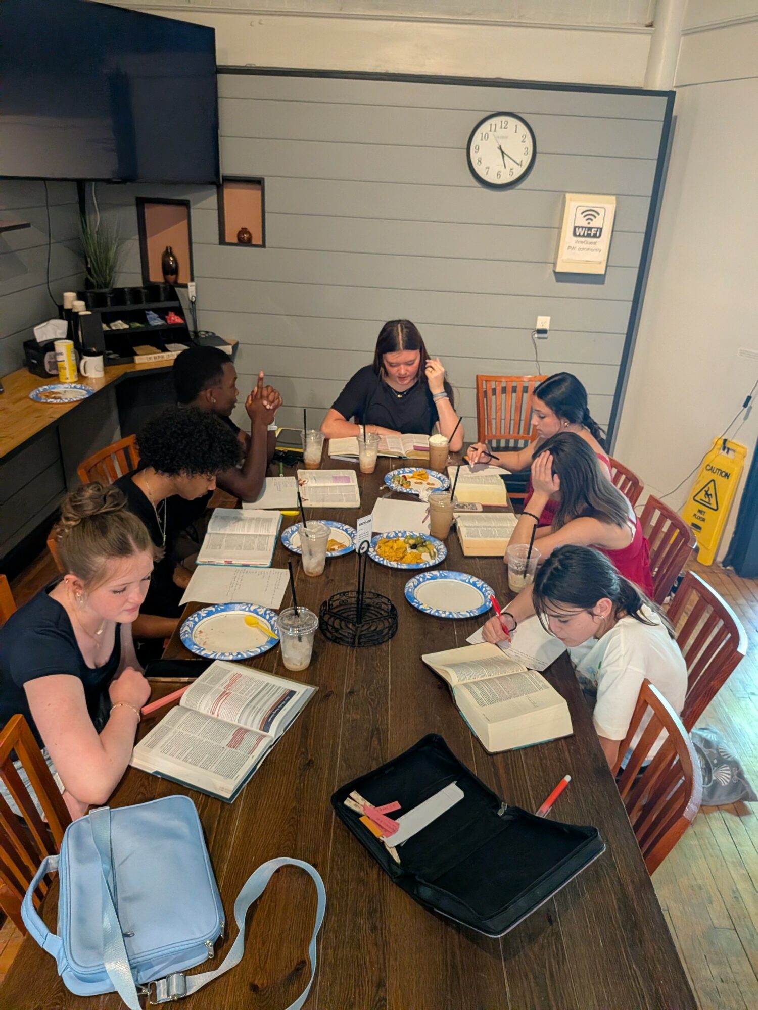 Six people sit around a wooden table reading books in a room with a clock and wall decorations.