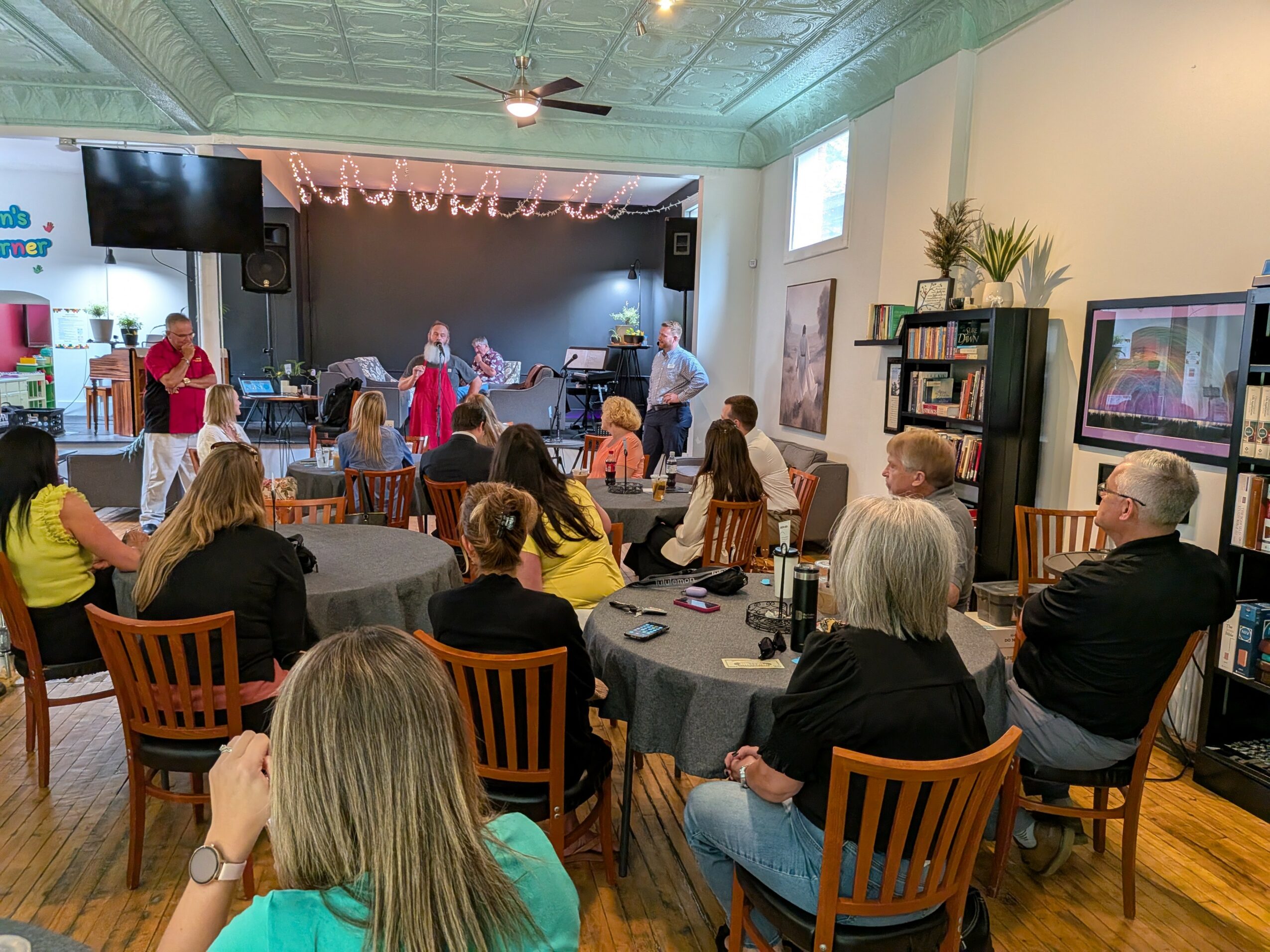 People watching a stage performance in a decorated room with tables and chairs, some watching TV screens, and a group on stage.