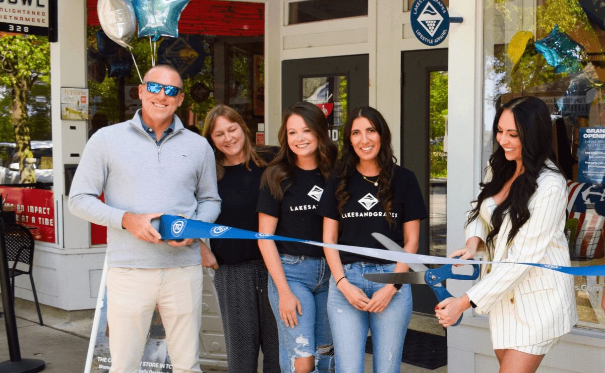 Group of five people at a ribbon-cutting event outside a store, smiling and holding scissors, with balloons and decorations.