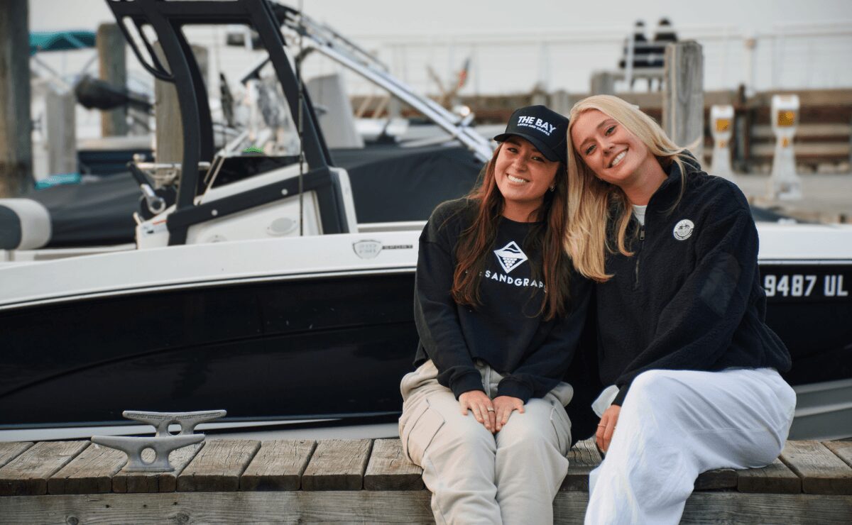 Two women sitting on a dock in front of a boat, smiling, with a marina in the background.