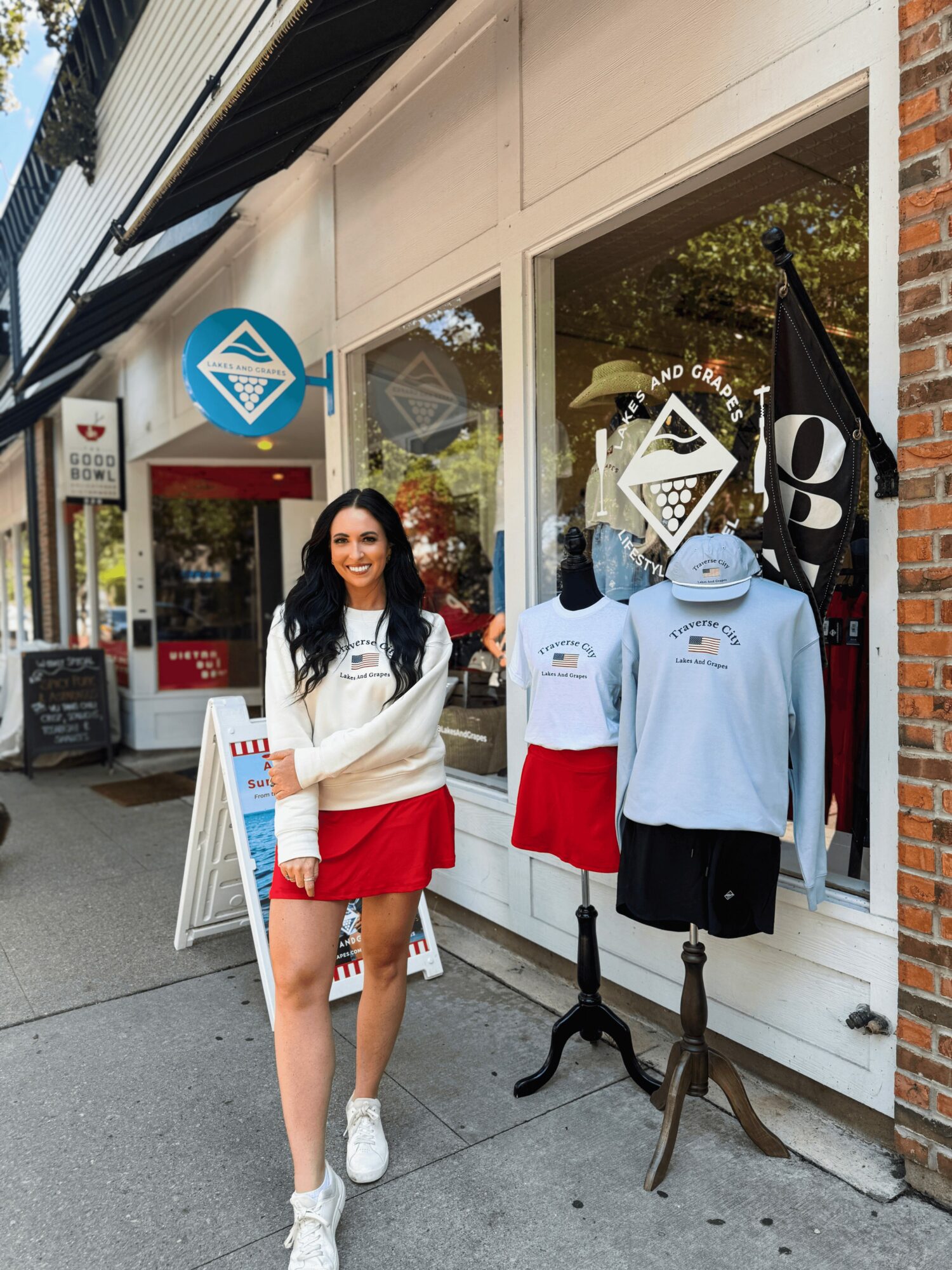 Young woman with long dark hair, wearing a white sweater and red skirt, standing outside a storefront with mannequins dressed in casual clothing.