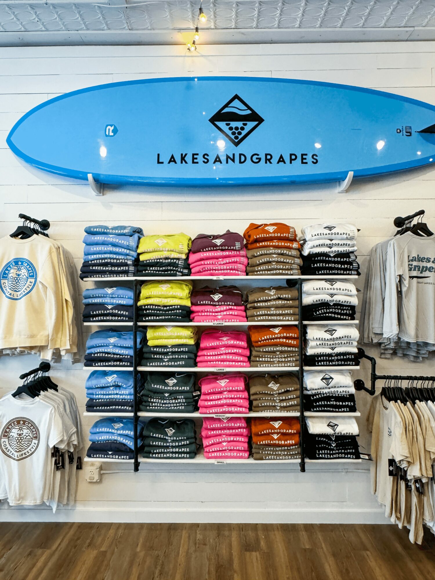 Blue paddleboard with 'LAKES AND GRAPES' logo mounted above clothing shelves in a store.