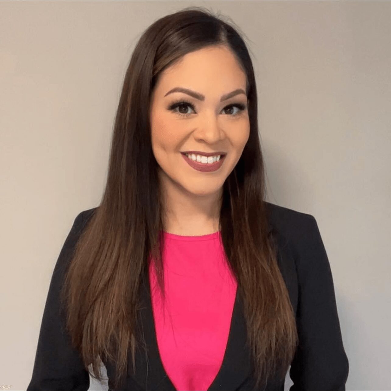 Woman with long brown hair smiling, wearing a black blazer and pink top, standing against a plain wall.
