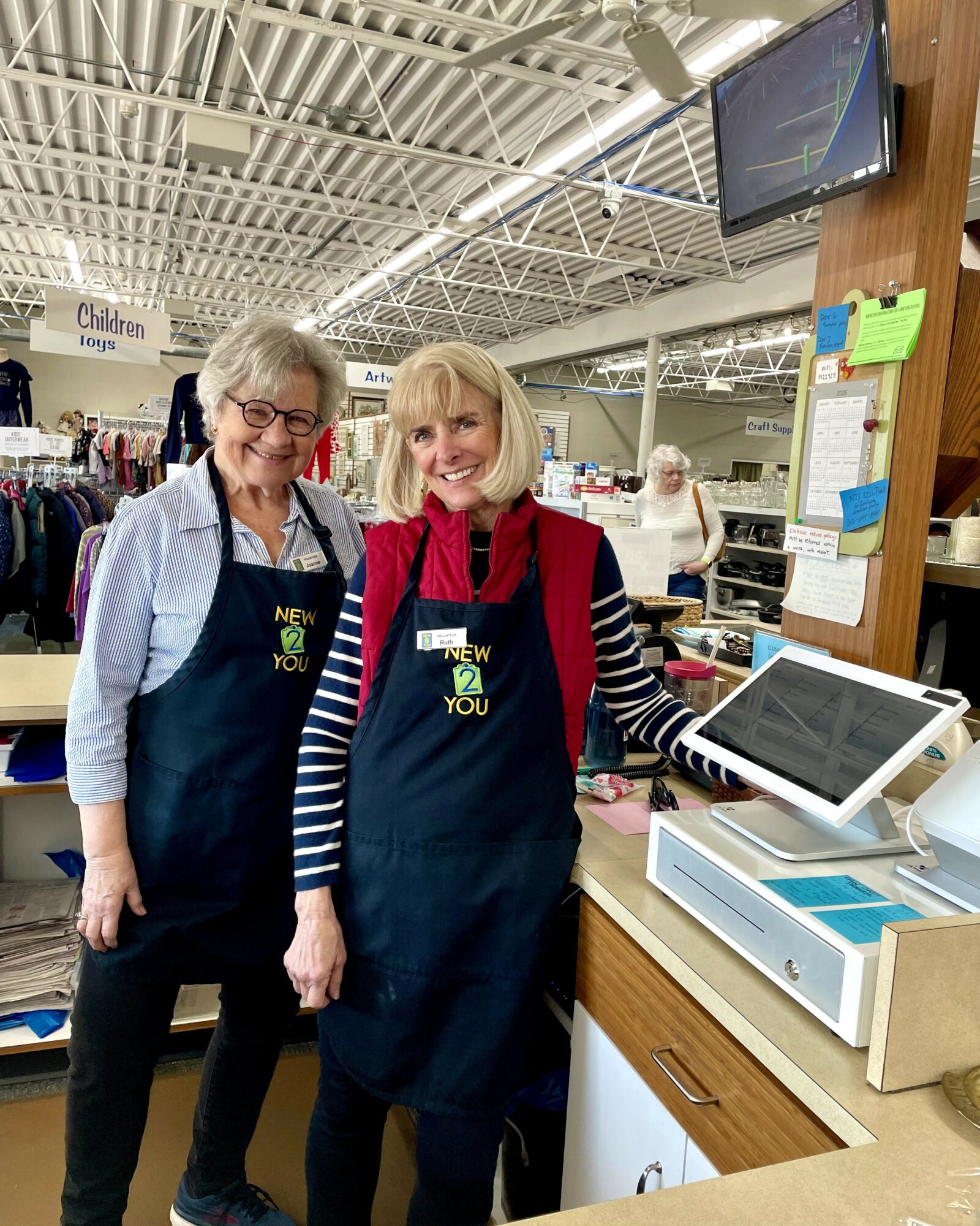 Two women wearing aprons stand behind a checkout counter in a store, smiling at the camera.
