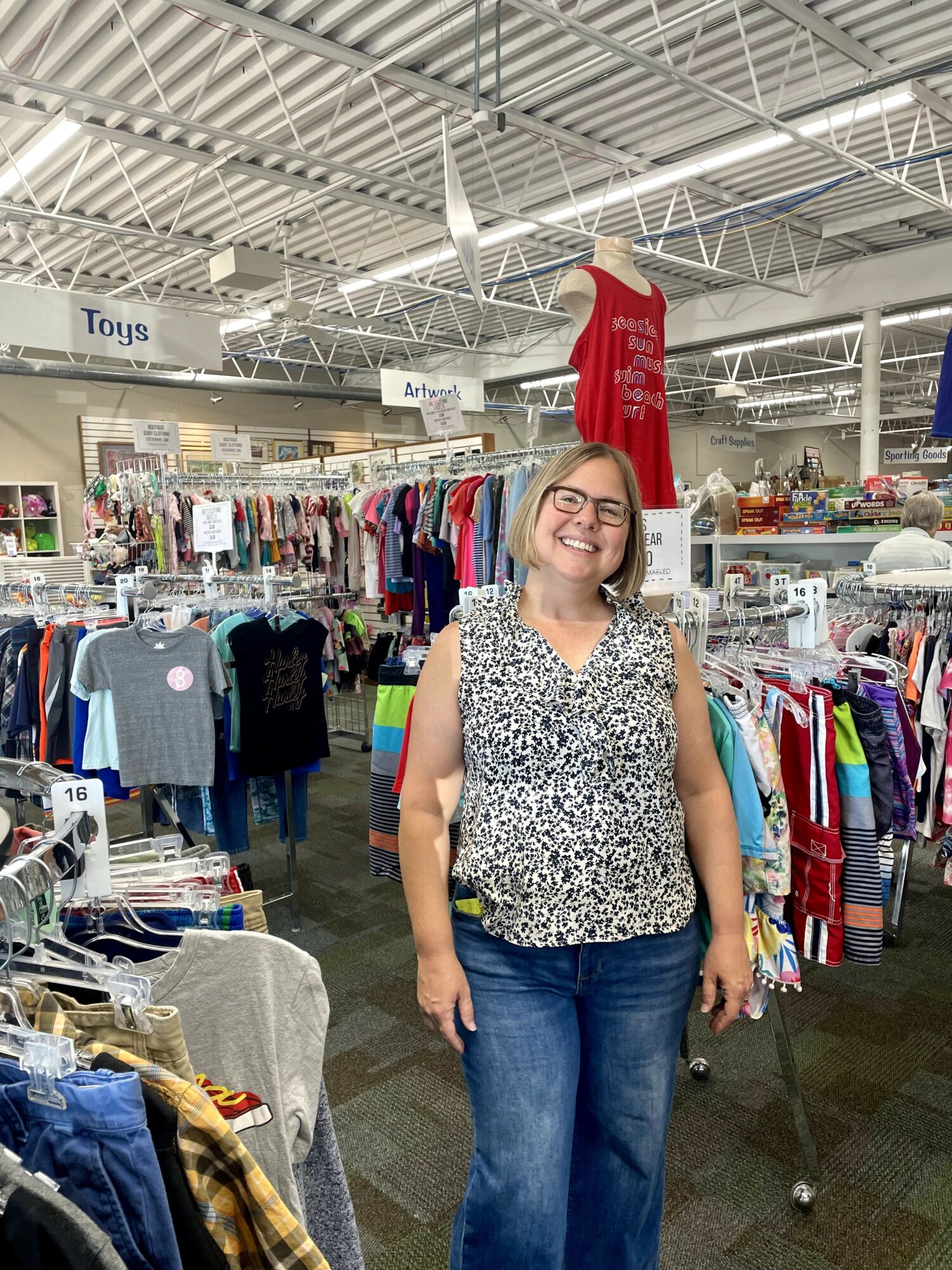 Smiling woman with glasses in a clothing store, surrounded by racks of clothes and toys, indoors with high ceiling.