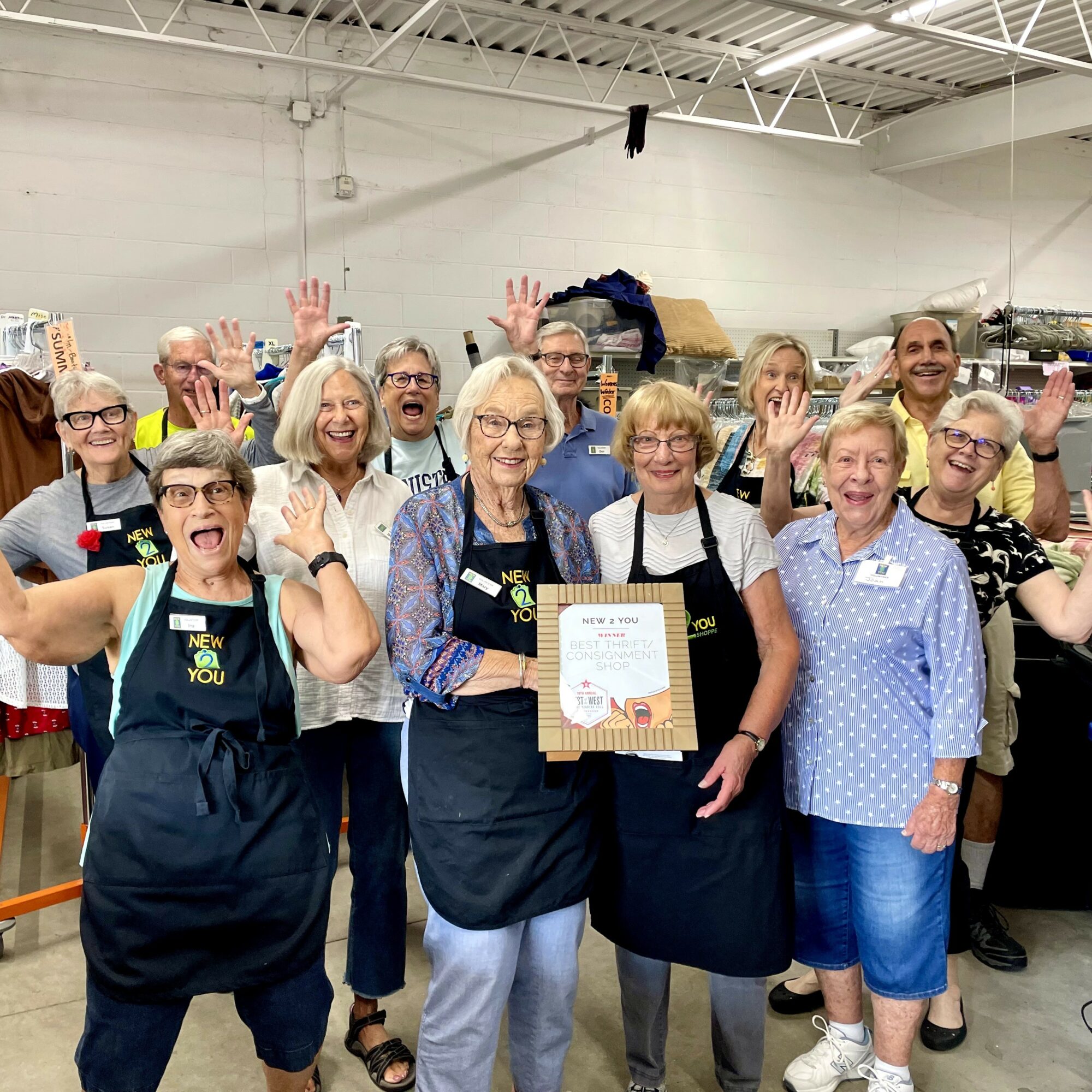 Group of smiling people in a warehouse, some waving, holding a certificate, wearing aprons and casual clothes.