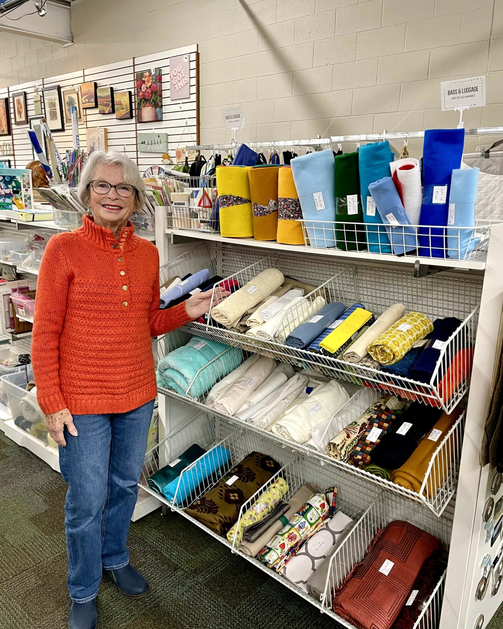Woman with glasses and orange sweater shopping for fabric or craft supplies in a store aisle.