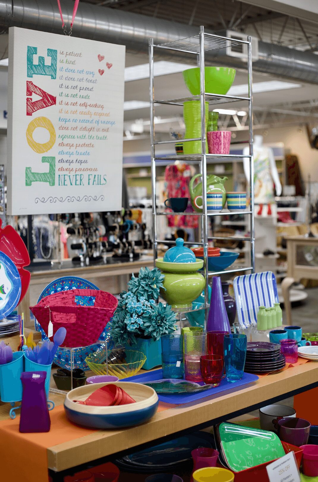 Display of colorful bowls, plates, and cups on a table with a shelving unit behind, in a store setting.