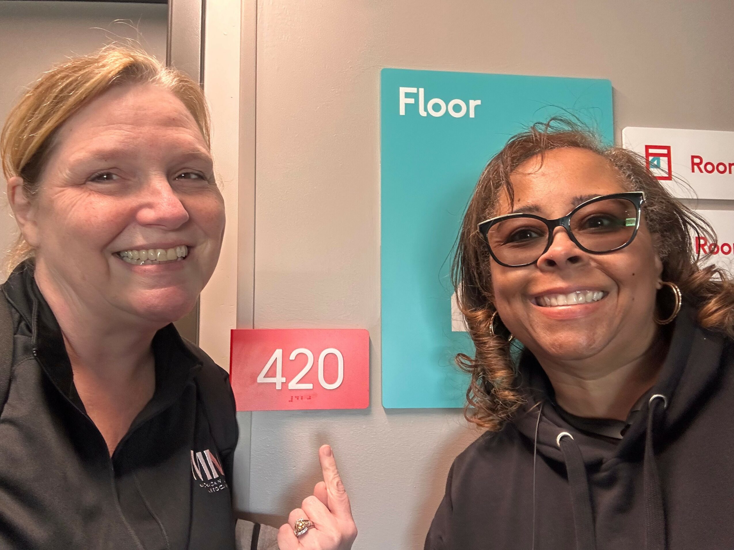 Two smiling women, one with glasses, pointing at a pink sign with '420', in front of a blue 'FLOOR' sign.