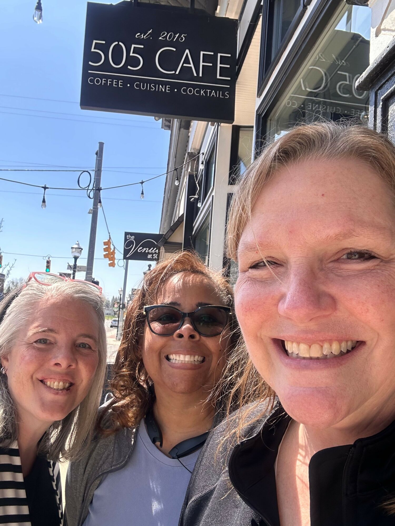 Three women smiling outside a cafe with a sign reading '505 CAFE' and a street scene in the background.