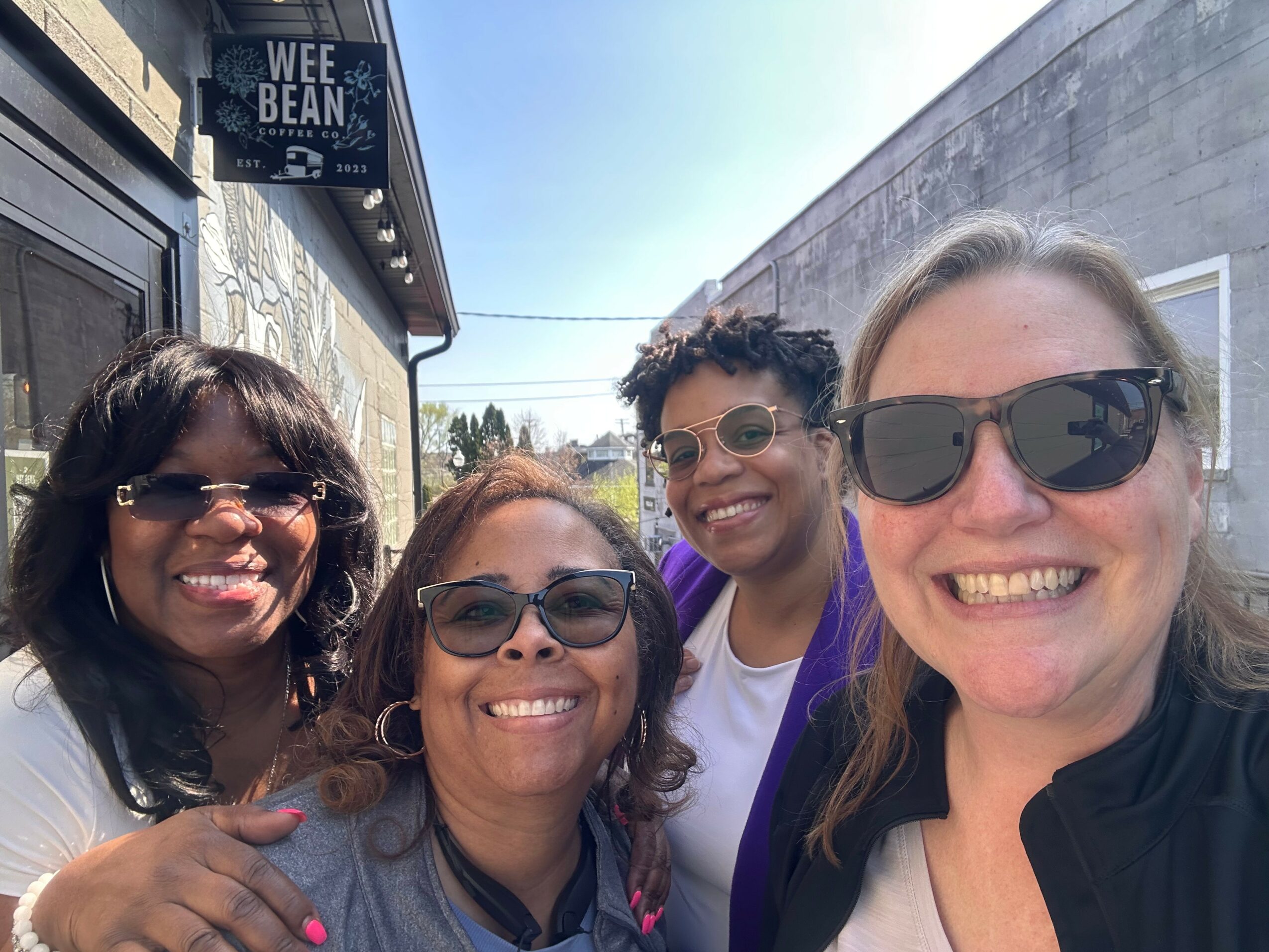 Four women smiling outdoors, wearing sunglasses, standing close together, with a building and sky in the background.