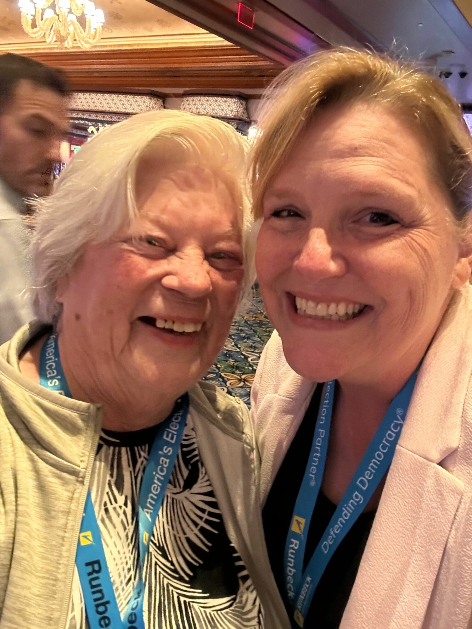 Two smiling women with blonde hair, wearing conference lanyards, close together indoors.