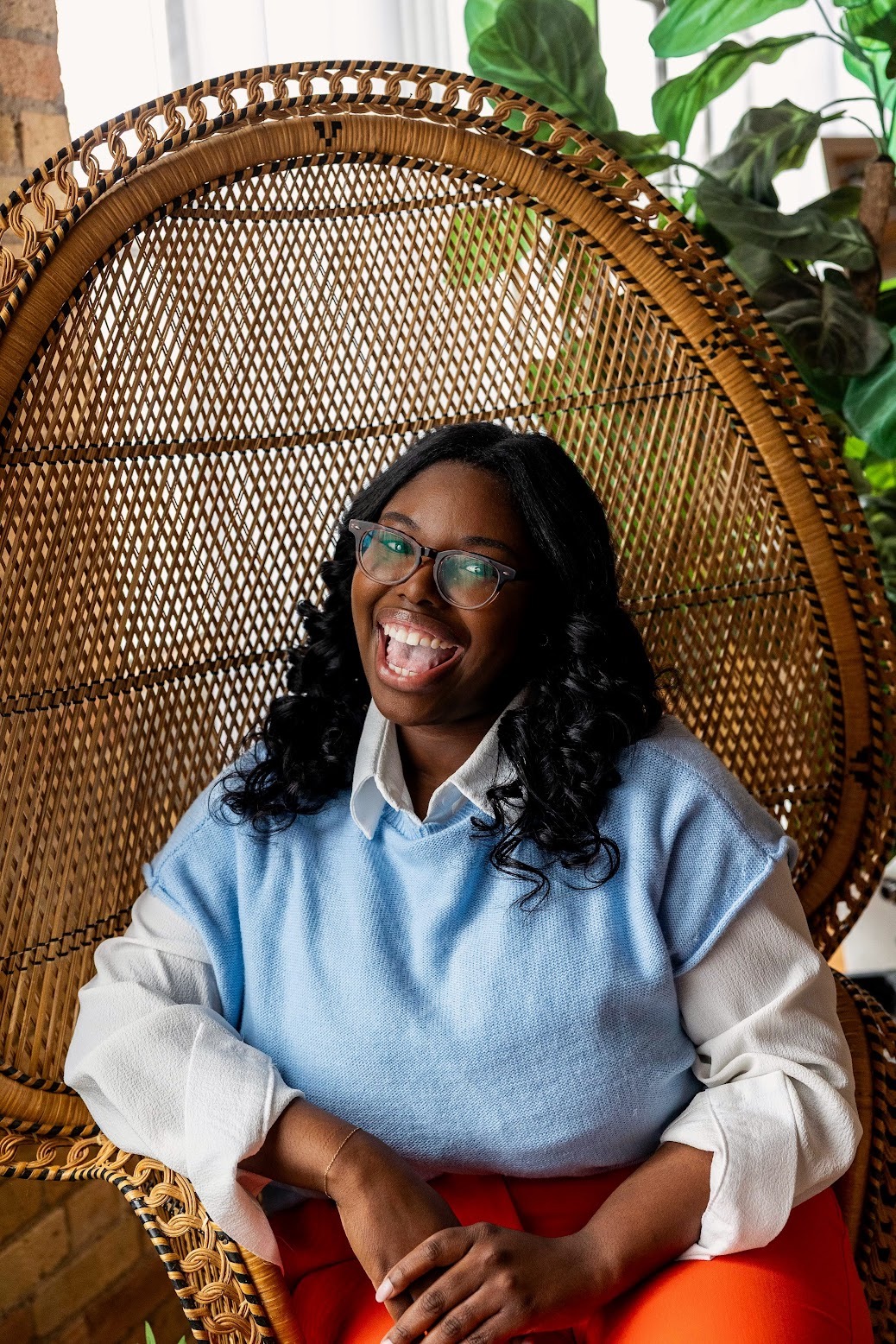 Woman with glasses smiling and sticking out tongue, sitting in a wicker chair with green plants in background.