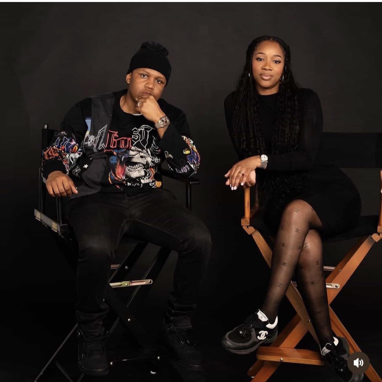 Two people sitting on chairs against a dark background, one with a beanie and colorful jacket, the other in a black dress with long braids.