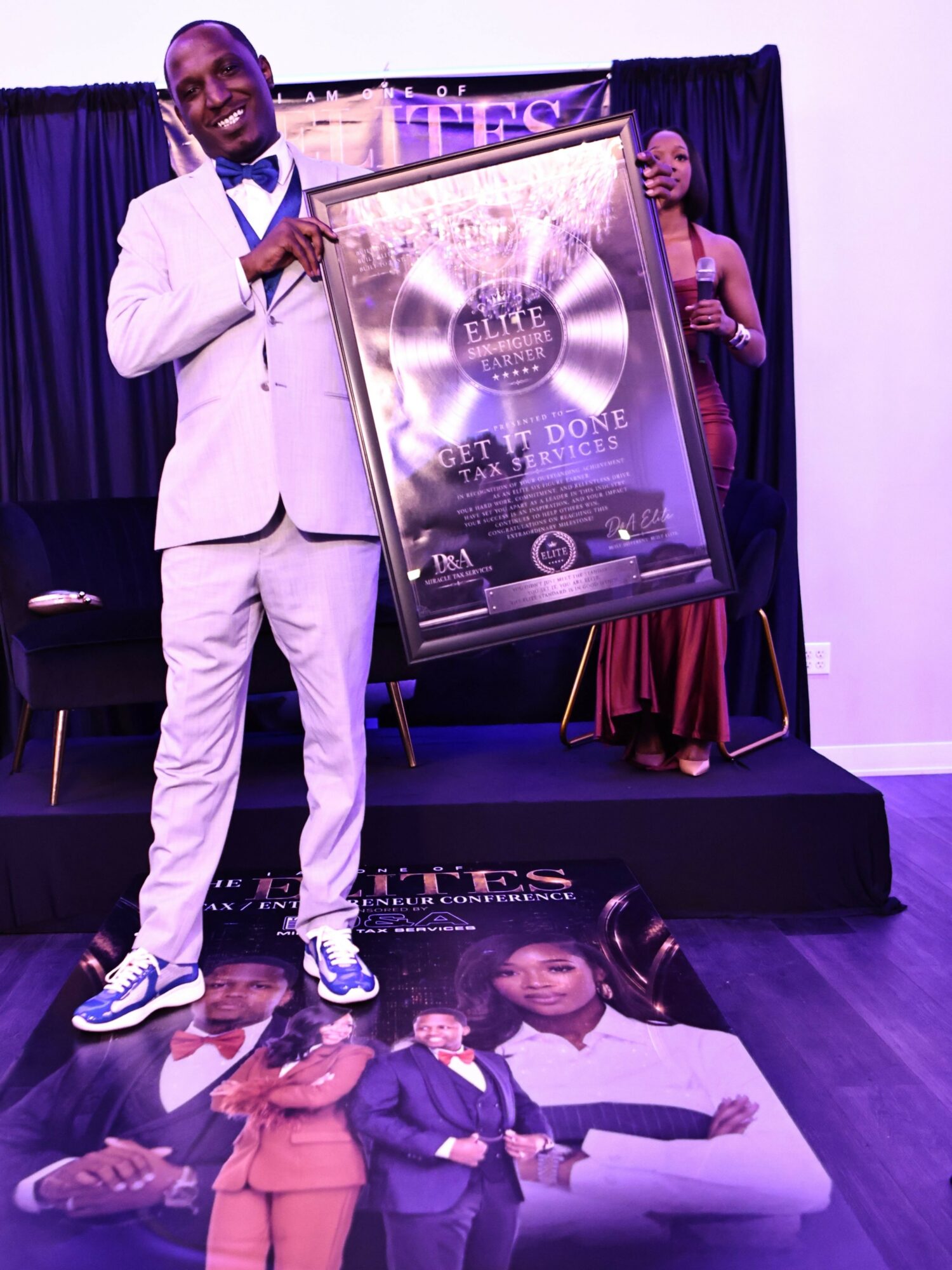 Man in white suit holding a framed award, woman in red dress standing behind, stage with black curtain backdrop.