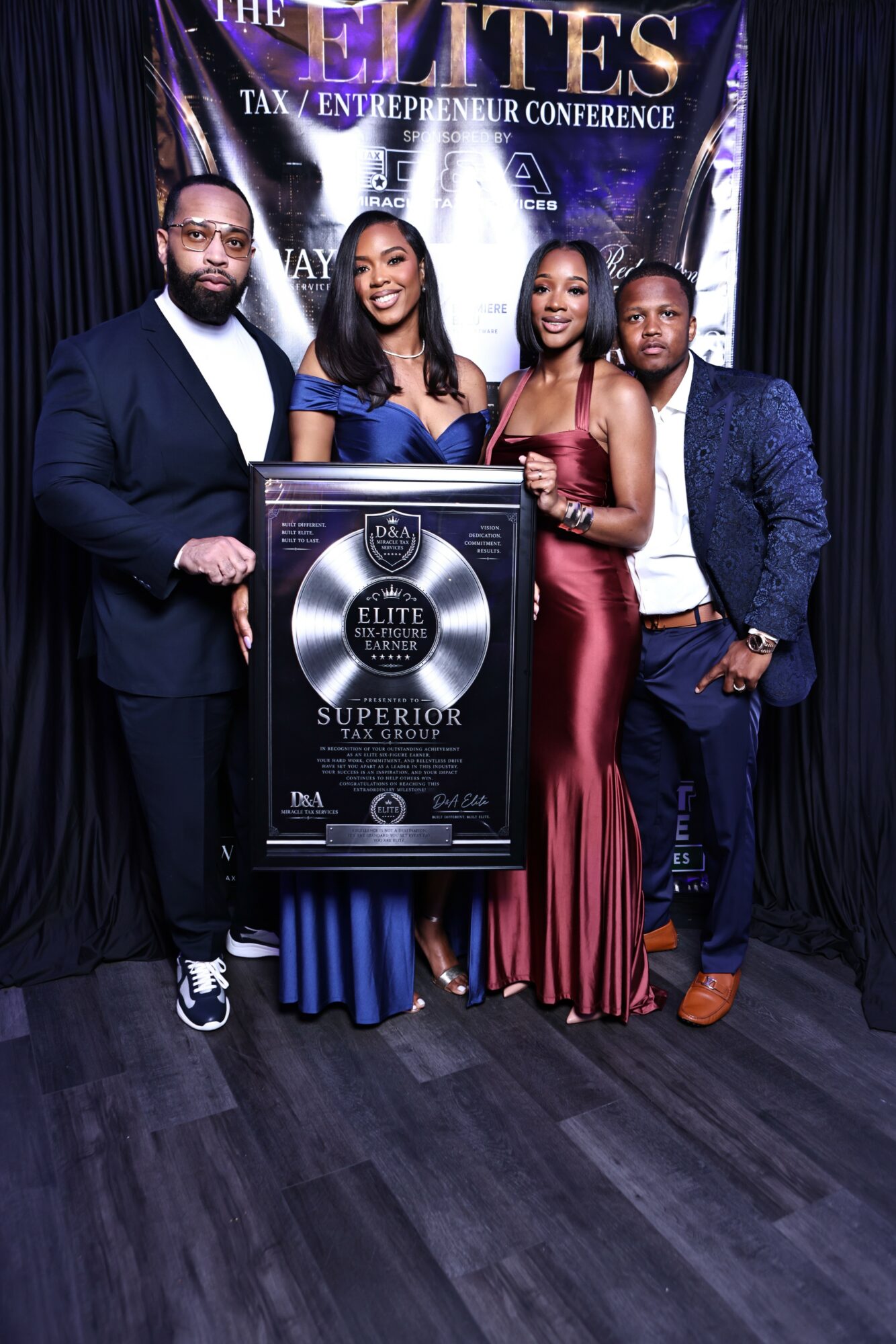 Four people standing together holding a large award plaque at a conference, with a banner in the background.