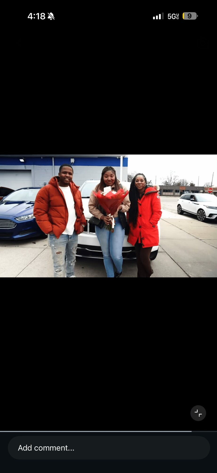 Three people standing outdoors in front of parked cars, one holding a bouquet of flowers, all wearing jackets.