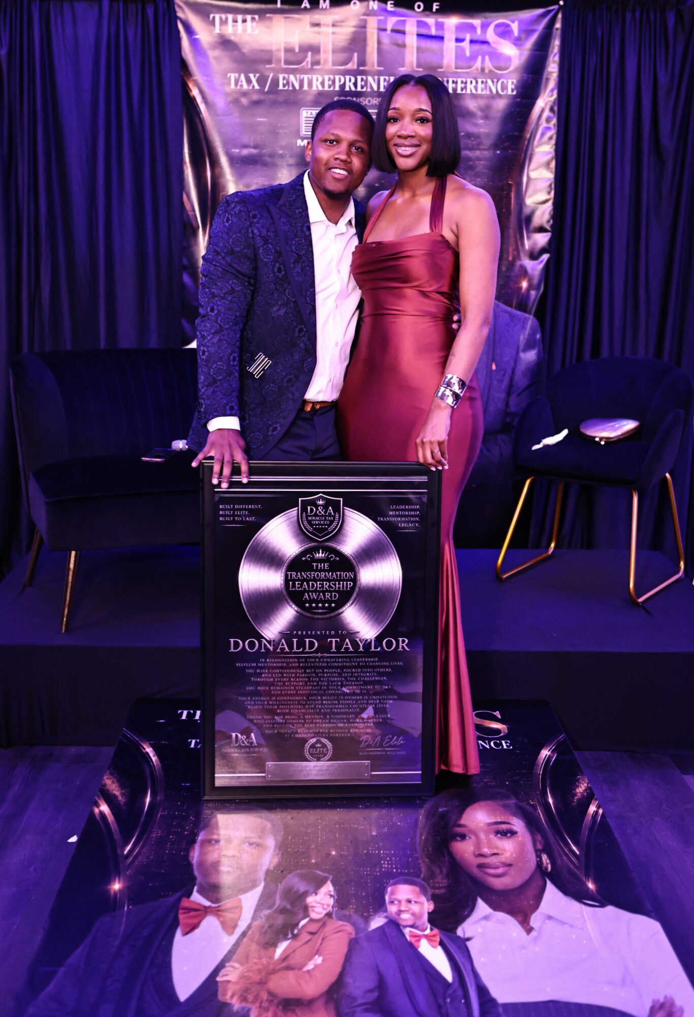 Two people stand on stage with a framed award in front, a woman in a red dress and a man in a suit, with a backdrop and chairs behind them.