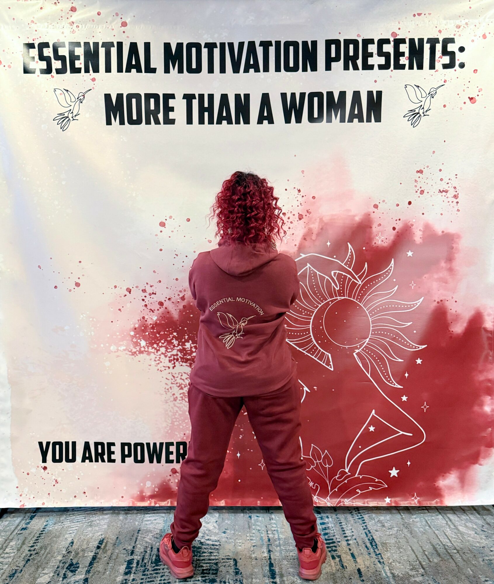 Person with curly hair facing a wall with motivational text and drawings, standing on a wooden floor. Red paint splatters on wall.